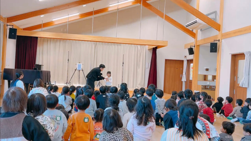 栃木県那須塩原市のひがしなす保育園でゲストの女の子と一緒にコップの水が消えるマジックを披露するCrystal Magician® Michelとそれを観る子どもたち