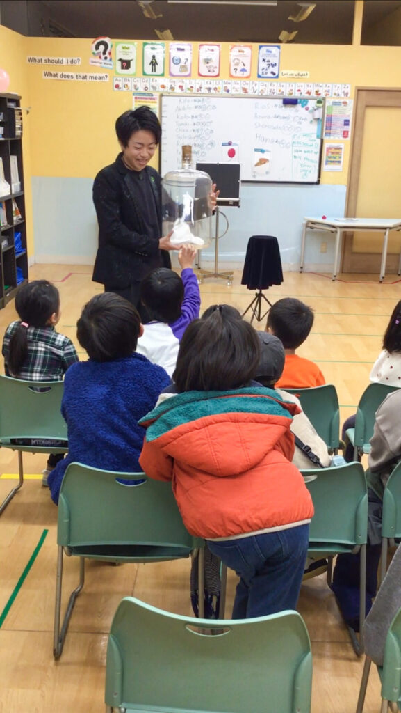 2026/1/5KidsDuo戸塚駅前のウィンタースクールイベントにてボトルの中でハンカチが踊るマジックを披露するCrystal Magician® Michelと夢中で見つめる子どもたち