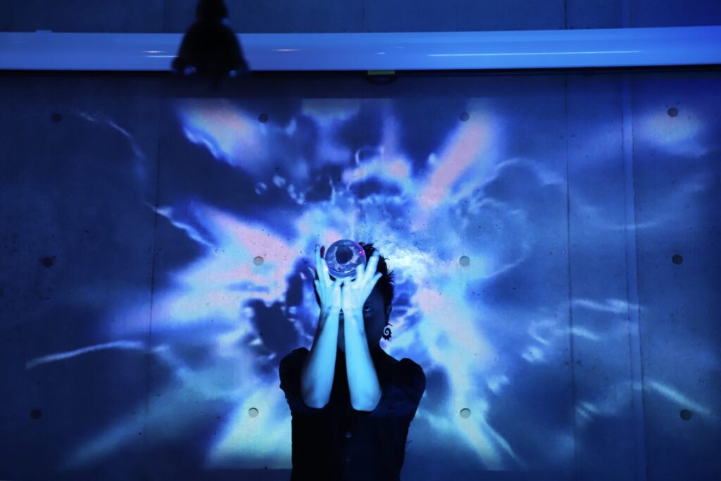 スクリーンに投影された稲妻の映像とLEDライトとともに水晶玉を使ったコンタクトジャグリングパフォーマンスを演じるCrystal Magician® Michel