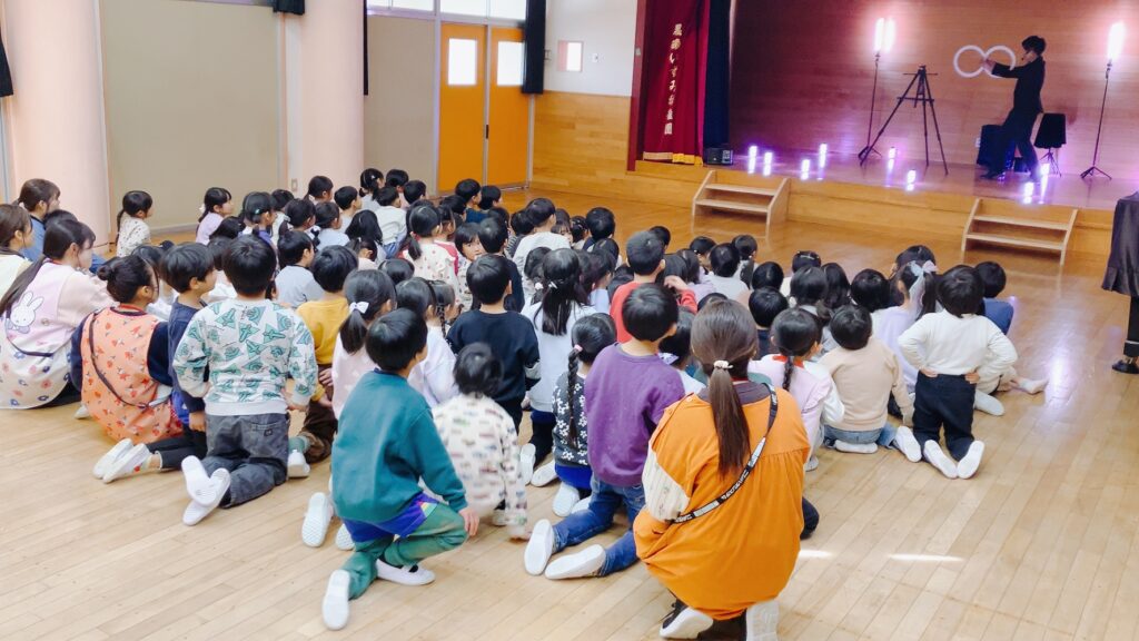 栃木県那須塩原市の黒磯いずみ幼稚園でLED演出とともに白いリングを使ったコンタクトジャグリングを演じるCrystal Magician® Michelとそれを観る子どもたち